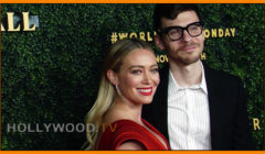 A man and a woman posing together at a red carpet event, with the woman wearing a red dress and the man in a black suit, standing in front of a hedge with logos.