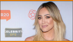 Hilary duff posing at a celebrity gps event, smiling with long blonde hair and subtle makeup, in front of a logo backdrop.