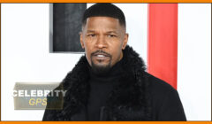 Man in black outfit and fur collar standing against a white and red background at a celebrity event.